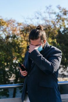 young businessman with long hair and beard, brown, laments because he receives bad news from his smartphone. He is outside near his offices during his work break. Gets bad coronavirus news from mobile phone.