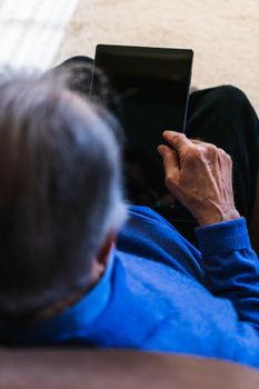 Grandfather with tablet in hands looking something. He is in a sofa, living room, home. Have a blue sweater. Upper view with natural light