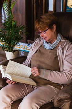 Caucasian old aged woman changing a page of book in a sofa. She is relaxing in her house while reading with purple glasses