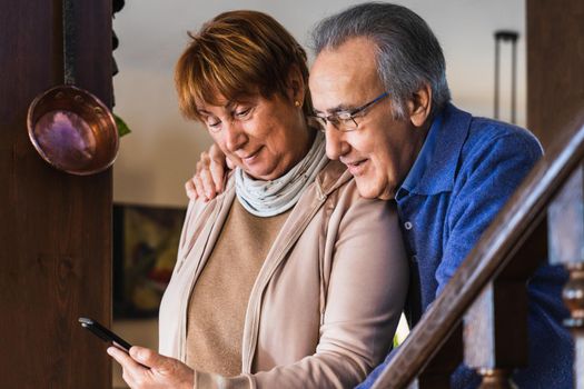 Grandparents looking smartphone in living room with natural light. Marriage together in home. She have the phone and he wear blue sweater and glasses. They are stand up in a rustic and traditional house