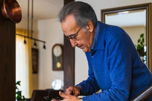 Old man looking smartphone stand up in home. He wear a blue sweater and glasses. Vertical shot of caucasian senior male with phone with natural light.