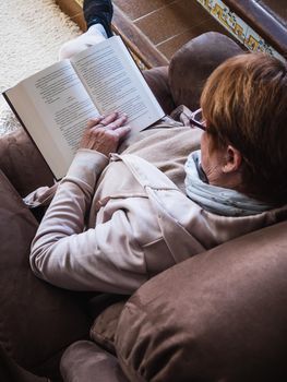 Old caucasian woman reading a book with purple glasses in sofa. Its a half size-back view with natural light