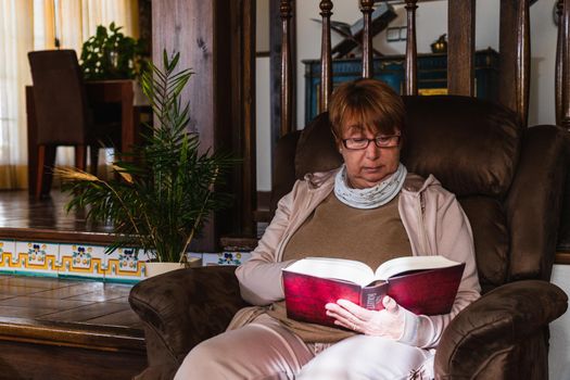 Old woman reading a red book in her sofa with purple glasses. She is in a sofa in her house with natural light. Its a rustic house