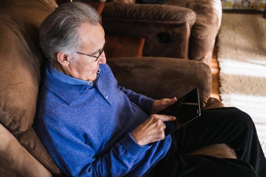 Grandfather man looking a tablet in living room. He is in a sofa and wear a blue sweater. He have glasses. Natural light in house