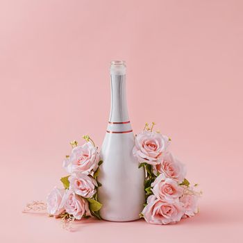 White bottle of champagne with garland of pink roses over a matching pink background with copy space for your greeting
