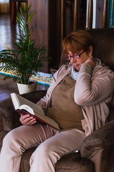 Grandmother reading a red book in a sofa while massage her neck. Its a half front-size detail view of her. She is in her house looking a book with natural light