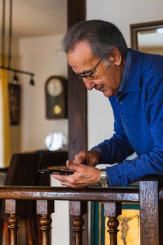Old man looking smartphone stand up in home. He wear a blue sweater and glasses. Vertical shot of caucasian senior male with phone with natural light.