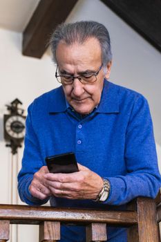 Old man looking smartphone stand up in home. He wear a blue sweater and glasses. Vertical shot of caucasian senior male with phone.