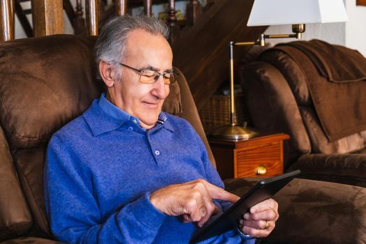 Grandfather man looking a tablet in living room. He is in a sofa and wear a blue sweater. He have glasses. Natural light in house