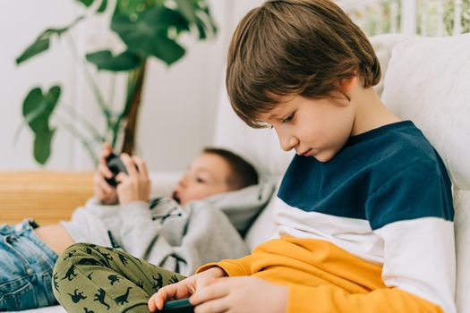 Cheerful two brothers boys playing online game, watching video on cellphone. Smiling kids using funny mobile apps, enjoying free leisure time at home on couch. Children texting together at phone.