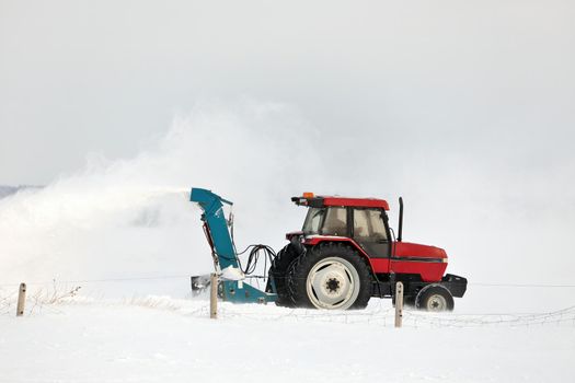 Red Tractor with snowblower attachment Snow Blowing a Driveway in a Rural Setting. It's windy and the blown snow creates a large plume of snow. High quality photo