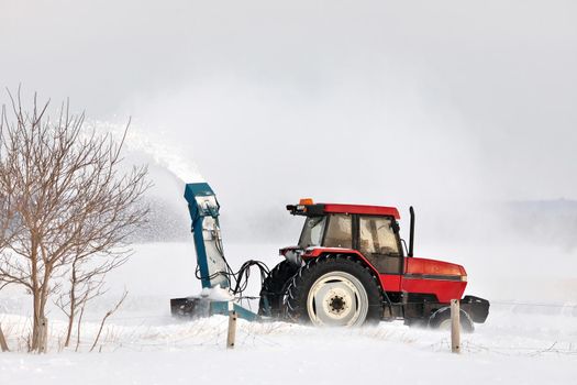 Red Tractor with snowblower attachment Snow Blowing a Driveway in a Rural Setting. It's windy and the blown snow creates a large plume of snow. High quality photo