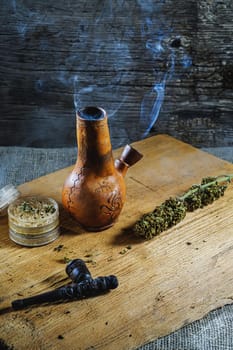 Old brown steamung bong and open cannabis grinder with chopped marijuana weed and medical cannabis bud in smoke by wooden desck and sackcloth. Vertical orientation