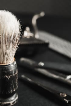 On a black surface are old barber tools. shaving brush vintage manual hair clipper, comb, razor, hairdressing scissors. black monochrome. Close-up. Barbershop background. contrast shadows. Vertical orientation.