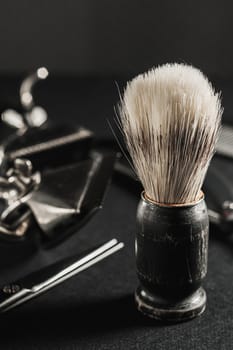 On a black surface are old barber tools. shaving brush vintage manual hair clipper, comb, razor, hairdressing scissors. black monochrome. Close-up. Barbershop background. contrast shadows. Vertical orientation.