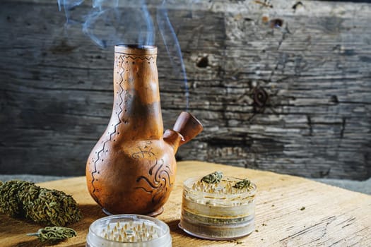 Old brown steamung bong and open cannabis grinder with chopped marijuana weed and medical cannabis bud in smoke by wooden desck and sackcloth. Horizontal orientation