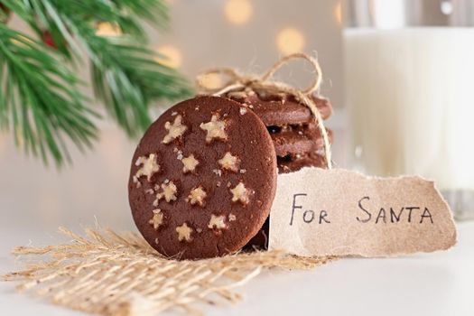 Santas cookies: a stack of round chocolate Christmas cookies with white sugar stars and salt tied with a rope, a glass of milk and a note, stand on a white table, under a spruce branch, shining garlands in the background