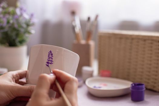 Close-up of female hands paint a sprig of lavender flower with a brush, on a white ceramic pot, with a brush. There are brushes, jars of paints and a palette, a wicker box on the table. Close-up.
