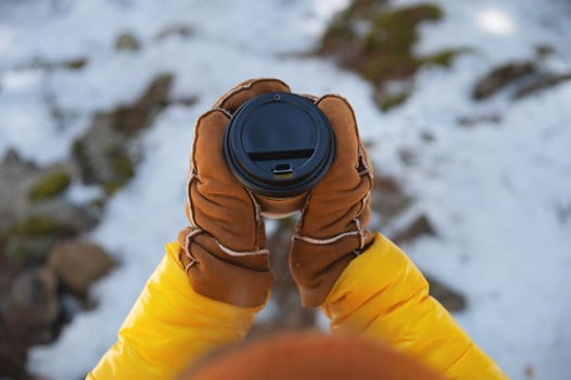 a paper cup with a black lid in hands in mittens, in nature against a background of snow