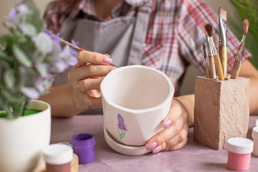 A hands of woman in a gray apron paints a white ceramic flower pot at the table in daylight. On the table is purple kraft paper, brushes, jars of paints. Close-up. No face