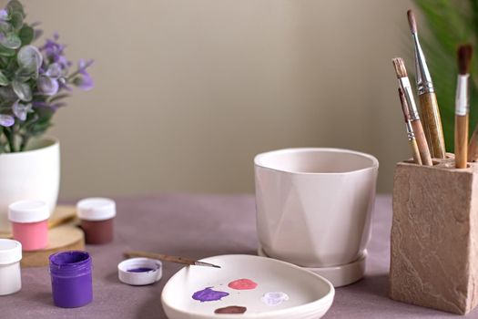 A still life with a white ceramic flower pot stands on a table on purple kraft paper, a palette, brushes in a stand, pink and purple paint cans. Copy space