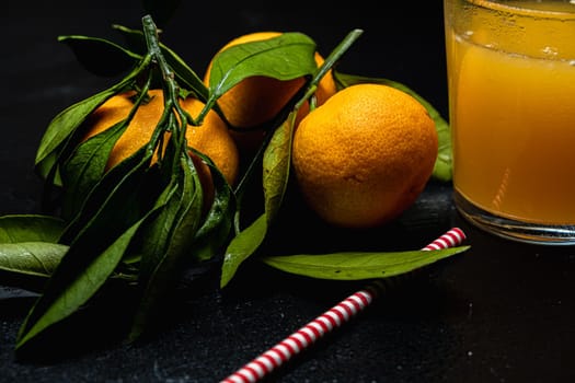 on a dark surface are three tangerines with leaves next to a glass of juice. Nearby lies a red and white striped straw. Horizontal orientation.