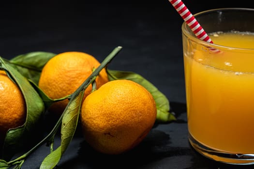 on a dark surface are three tangerines with leaves next to a glass of juice. Horizontal orientation.