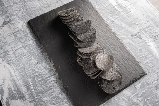 a lot of black chips with spices on a slate dish that lies on a concrete texture.