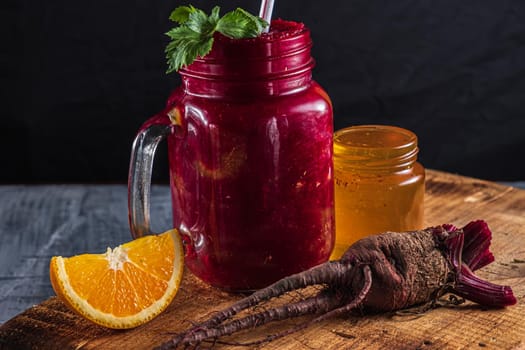 Smoothie consists of beets, orange, celery in a mason jar. There is a jar of honey next to it, a beet root, a piece of pumpkin and pieces of orange. on a thick wooden board. horizontal orientation.