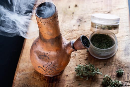 Old bong and open cannabis grinder with chopped weed and marijuana in smoke are located on the board. Wooden background. Horizontal composition.