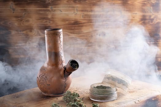 Old bong and open cannabis grinder with chopped weed and marijuana in smoke are located on the board. Wooden background. Horizontal composition.