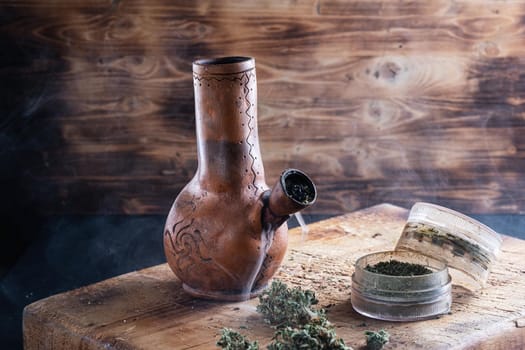 Old bong and open cannabis grinder with chopped weed and marijuana in smoke are located on the board. Wooden background. Horizontal composition.