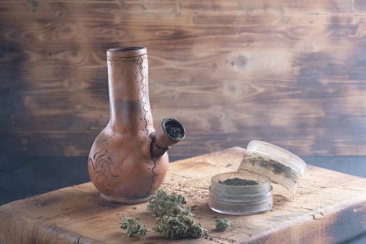 Old bong and open cannabis grinder with chopped weed and marijuana in smoke are located on the board. Wooden background. Horizontal composition.