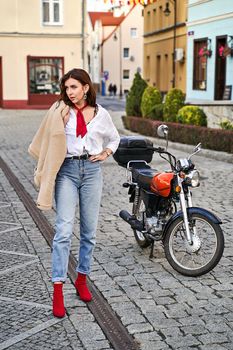 Young beautiful woman wearing red knitted fabric block heel sock shoes, blue denim jeans pants and teddy jacket coat posing near a motorcycle on a street