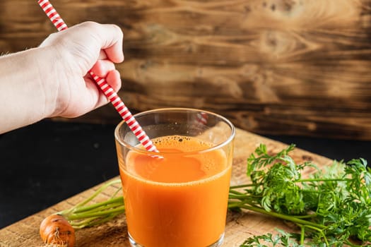 one litle ugly carrot. Glass with freshly squeezed carrot juice. one ugly carrot with green leaves. plastic free. cardboard tube. Wood background. horizontal orentecion. Children's hand reaches for a straw