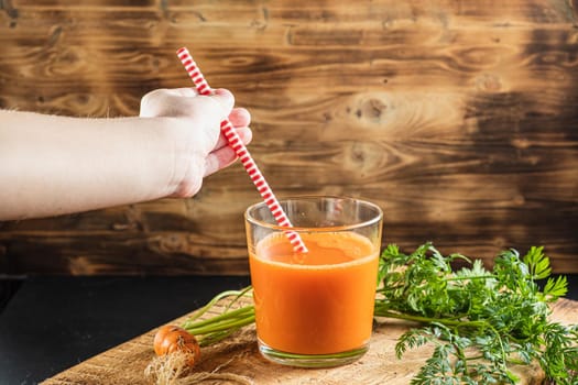one litle ugly carrot. Glass with freshly squeezed carrot juice. one ugly carrot with green leaves. plastic free. cardboard tube. Wood background. horizontal orentecion. Children's hand reaches for a straw