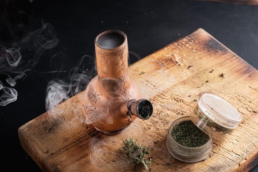 Old bong and open cannabis grinder with chopped weed and marijuana in smoke are located on the board. Wooden background. Horizontal composition.