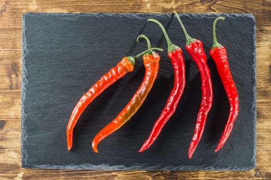 five red hot peppers lie on a slate and a wooden surface