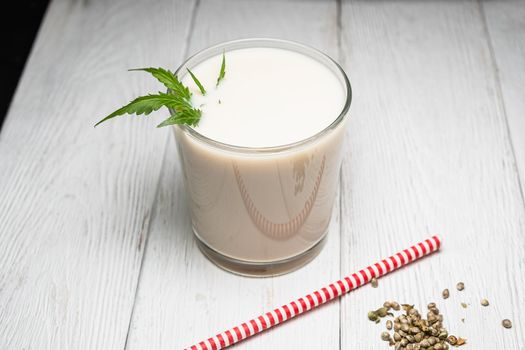 Hemp milk is poured into a glass. On the surface is a cannabis leaf. Nearby marijuana seeds. On the white old weathered boards. Near the red and white non-plastic straw High key