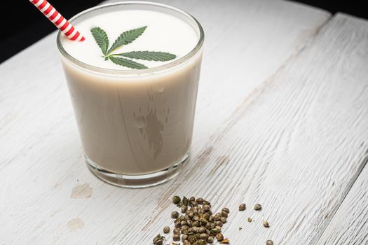 Hemp milk is poured into a glass. On the surface is a cannabis leaf. Nearby marijuana seeds. On the white old weathered boards. In a glass, red and white non-plastic straw.