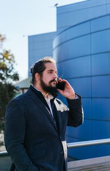 Young businessman with long hair and beard calling during his work break, on the street next to the offices. Vertical photo on a sunny and clear day.