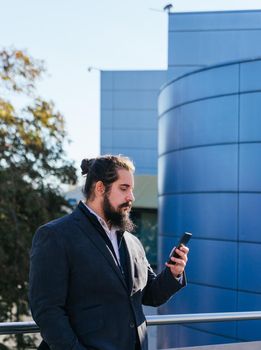 Young businessman with long hair and beard looking smartphone during his work break, on the street next to the offices. Vertical photo on a sunny and clear day.