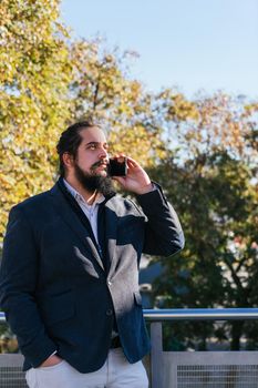 Young businessman with long hair and beard calling during his work break, on the street next to the offices. Vertical photo on a sunny and clear day.