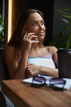 young asian pretty woman sitting at table of coffee shop, calling by smartphone. beautiful lady talking by cell phone at cafe, holding laptop. modern communication technology, distant work, remote job