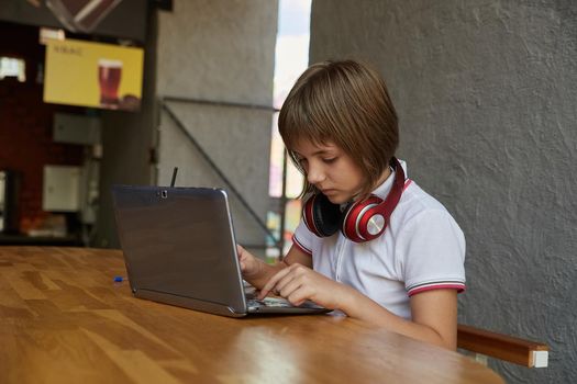 schoolgirl sits outdoors, types on keyboard of laptop. little caucasian girl sitting at table at veranda of cafe, studying with computer, doing homework. distant learning, modern online education