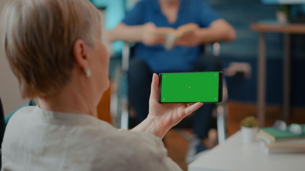 Female pensioner looking at horizontal green screen on mobile phone. Old woman with crutches holding smartphone with blank chroma key and isolated copy space background. Mock up template