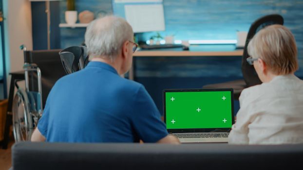 Senior couple using laptop with green screen in living room. Elder man and woman looking at blank chroma key with isolated copy space background and mock up template on computer display.