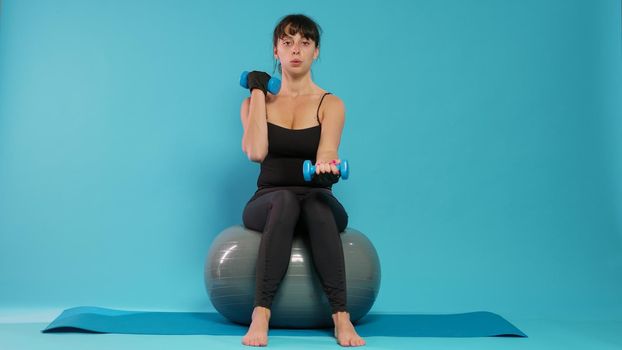 Fit person using dumbbells to lift weights on fitness toning ball, doing physical exercise to train body muscles. Athletic woman using pilates equipment on yoga mat in front of camera.