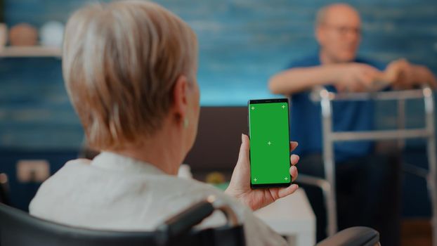 Senior woman in wheelchair holding smartphone with green screen at home. Retired person with chronic disability using isolated copy space with blank mock up and chroma key background on display.