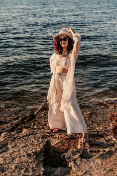 A woman in a white pantsuit and hat is standing on the beach enjoying the sea. Happy summer holidays.